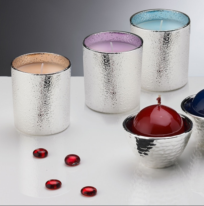 Candele profumate in bicchieri argentati con riflessi textured, diverse colorazioni di cera e piccoli gemme rosse decorative su tavola bianca.