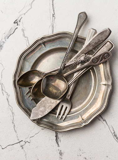 Set di posate d'argento usurate adagiate su un piatto decorato: cucchiai, forchetta e coltello con manici ornati su sfondo marmoreo.