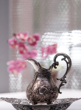 Antico boccale argentato decorato con motivi floreali, manico ricurvo, sfondo sfocato con fiori rosa.