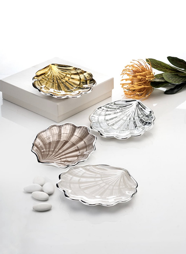 Set di piatti a forma di conchiglia in metallo rifinito, in oro, argento e bronzo su piano bianco con fiore giallo e nastri decorativi