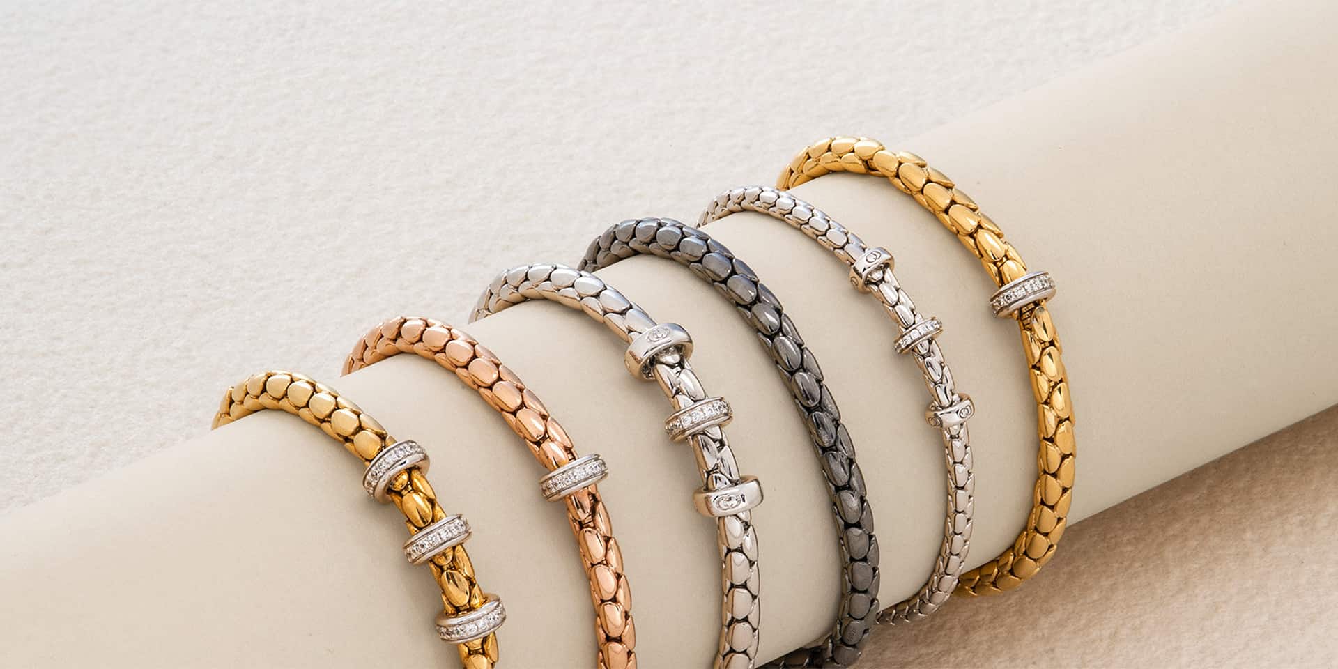 Bracciali a intreccio in tonalit&agrave; oro, rame, argento e grigio, con charm brillanti, indossati su polso chiaro