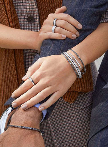 Mano con orologi e bracciali metallici, primo piano su anello e accessori su sfondo tessuti e pelle, stile sofisticato e elegante.