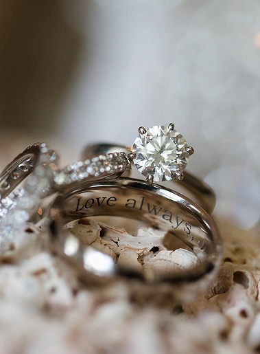 Anello con pietra brillante al centro e incisione 'Love always' all'interno, brillantini e anelli sfumati sullo sfondo.