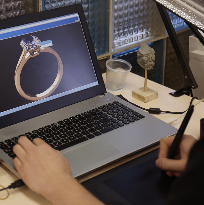 Immagine di una persona che lavora su un laptop, progetti di gioielli e oreficeria sullo schermo, con strumenti di modellazione e una piccola statua in primo piano.