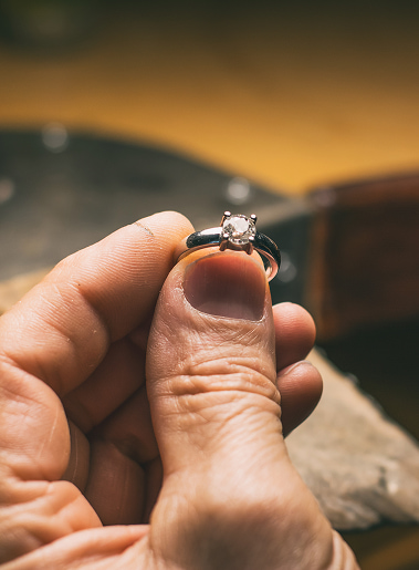 Anello di fidanzamento con brillantino tenuto tra le dita, anteprima di gioielleria con sfondo sfumato.