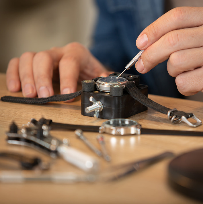 Riparazione orologio da tasca: mani al lavoro con utensili su cassa, cinturino e ingranaggi sparsi su tavolo in legno.