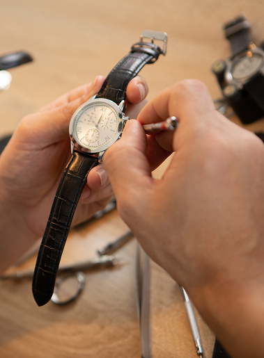 Orologiaio aggiusta orologio da polso con cinturino nero, dettaglio sul quadrante e strumenti di riparazione sul tavolo